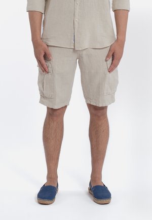 Beige linnen cargoshorts met twee zijzakken, gecombineerd met blauwe espadrilles met een geweven jute zool. De stof heeft een natuurlijke textuur.