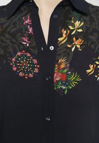 Camicia nera con bottoni, caratterizzata da ricami floreali colorati, che includono vivaci tonalità di verde, rosa e arancione, con colletto classico e una texture liscia.