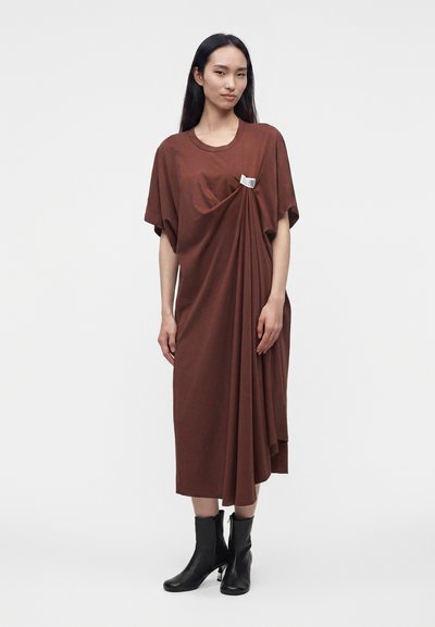 Γυναίκα με μακριά σκουρόχρωμα μαλλιά φοράει ένα φαρδύ καφέ midi φόρεμα με πλάγια draping και μαύρα μποτάκια αστραγάλου, στέκεται μπροστά σε απλό ανοιχτό φόντο.