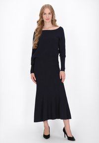 Rochie tricotată bleumarin cu mâneci lungi și decolteu off-the-shoulder, având un corset ajustat și un tiv evazat, asortată cu pantofi cu toc negri.