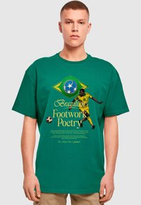 Vihreä puuvillapaita, jossa on jalkapalloilijan graafinen kuva. Tekstissä lukee "Footwork Poetry" ja brasilialaisia symboleja. Lyhythiha, rento malli.