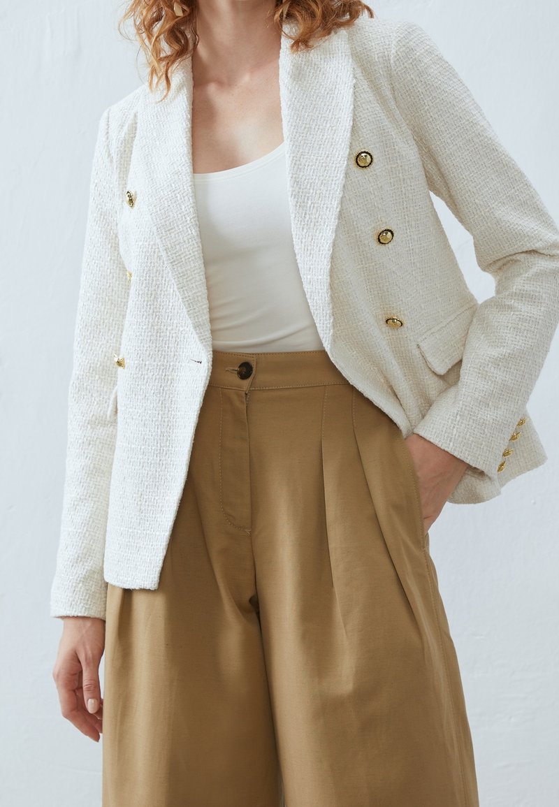 Femme portant un blazer blanc texturé avec des boutons dorés, un haut blanc et un pantalon kaki taille haute à jambes larges, une main dans la poche.