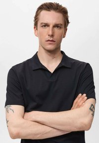 Svart polotröja med krage, korta ärmar och en texturerad tyg. Visar tatueringar på modeller som har armarna i kors.