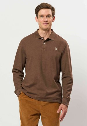 Jeune homme portant un polo marron à manches longues et un pantalon en velours côtelé marron clair, debout avec une main dans la poche sur un fond uni.