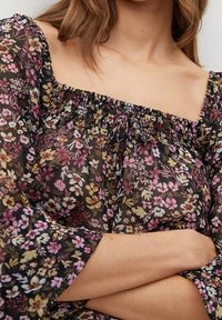 Kwiatowa bluzka z przezroczystego czarnego materiału, z kwadratowym dekoltem, marszczonym brzegiem i długimi rękawami ze elastycznymi mankietami. Akcenty w różowe, żółte i zielone kwiaty.