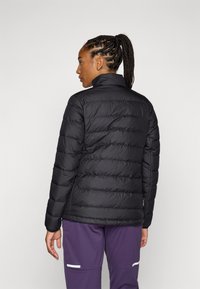 Zwarte pufferjack met een hoge kraag, doorgestikt ontwerp en elastische manchetten, gecombineerd met paarse broek met witte accenten.