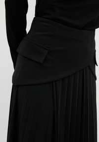 Jupe plissée noire avec une taille structurée, dotée d'une poche latérale et d'une texture lisse. Le design comprend un aspect superposé en haut.