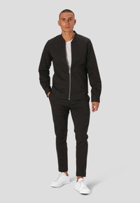 Črna zip-up jakna z okroglim ovratnikom, v kombinaciji s črnimi oprijetimi hlačami in belimi supergami. S sleek, minimalističnim dizajnom in mehkobo.