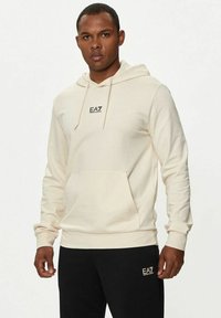 Felpa con cappuccio crema, dotata di tasca frontale, polsini a coste e un piccolo logo nero sul petto. Abbinata a pantaloni neri con logo.