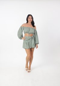 Mujer con cabello largo y oscuro, vestida con un top corto verde con estampado floral y hombros descubiertos y falda a juego, con sandalias blancas de tacón, de pie sobre un fondo blanco.