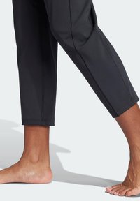 Zwarte cropped broek met een gladde textuur en contrasterende naden, met een zichtbare enkel-detail en een blote voet naast de stof.