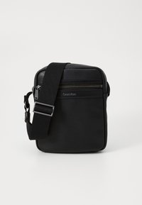 Schwarze Leder-Crossbody-Tasche mit strukturierten Oberflächen, verstellbarem Riemen, vorderer Reißverschlusstasche und subtiler Markenkennzeichnung an der Seite.