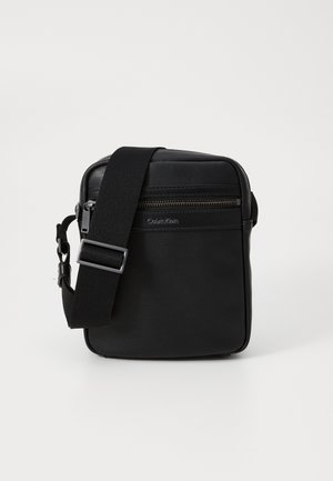 Sac en cuir noir à bandoulière avec une surface texturée, une sangle ajustable, une poche zippée à l'avant et un branding subtil sur le côté.
