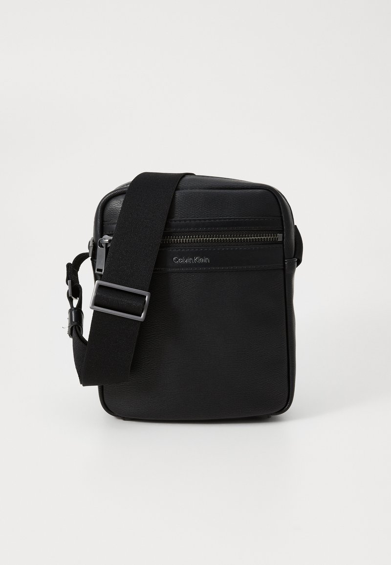 Schwarze Leder-Crossbody-Tasche mit strukturierten Oberflächen, verstellbarem Riemen, vorderer Reißverschlusstasche und subtiler Markenkennzeichnung an der Seite.