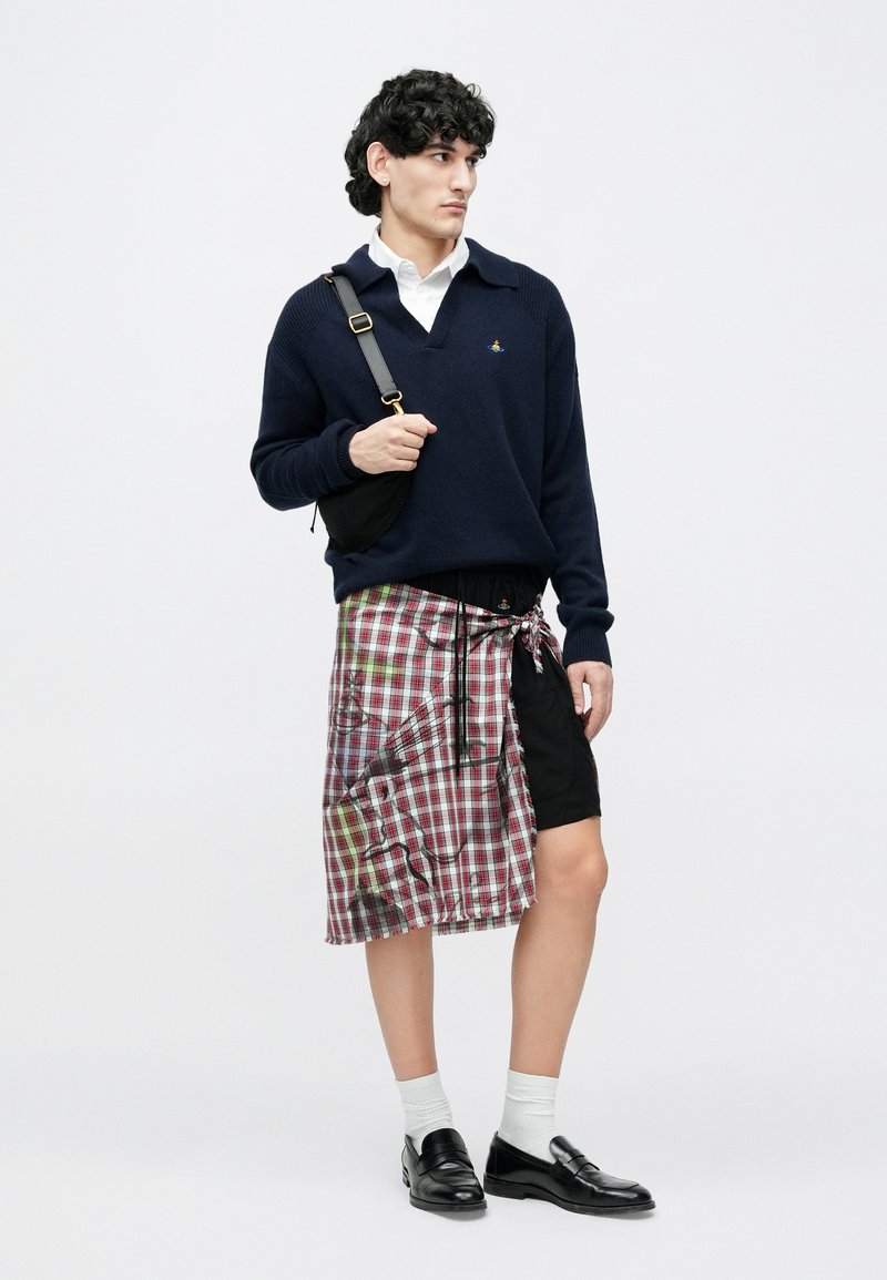 Marineblå sweater, hvid skjorte med krave, ternet omslagsnederdel i nuancer af rød og grøn, sorte shorts under, sorte loafers, båret med hvide sokker.