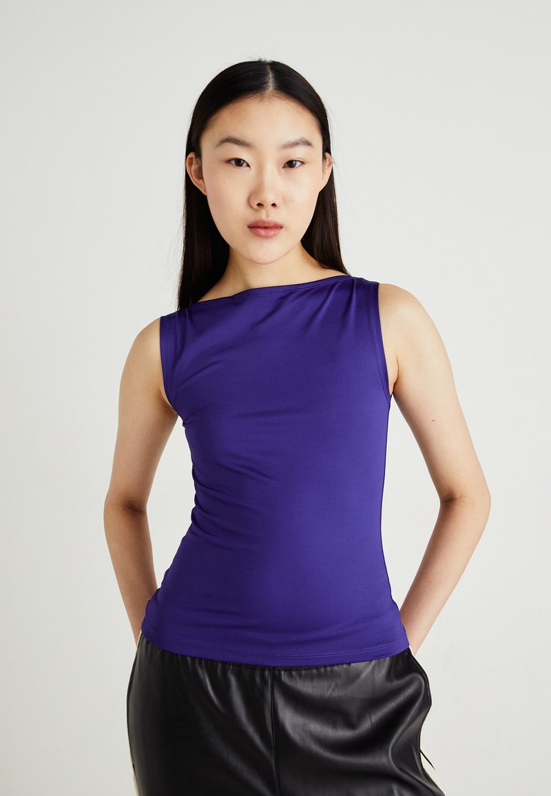 Haut sans manches violet en tissu lisse et extensible ; silhouette ajustée ; décolleté asymétrique ; associé à un pantalon en cuir noir.