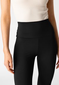 Vysoké černé legíny vyrobené z ribbrovaného materiálu s hladkým povrchem, s širokým páskem a přiléhavým střihem na nohách.