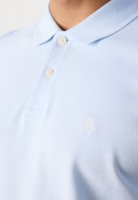 Gros plan sur une personne portant un polo bleu clair avec deux boutons fermés et une broderie blanche "M O P" sur la poitrine.