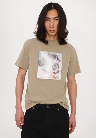 Camiseta de algodón beige con un estampado gráfico cuadrado de diseño abstracto. Mangas cortas y cuello redondo. Pantalones negros texturizados.