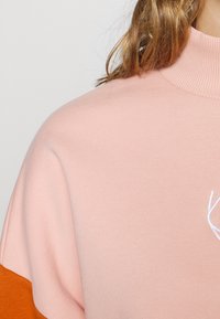 Felpa rosa con colletto alto a coste e dettaglio arancione. Presenta una texture morbida e un logo ricamato sulla zona del petto.