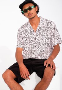 Kurze, knopflose Hemd mit rosa Grundfarbe und schwarzem abstraktem Muster. Kombiniert mit schwarzen Cordshorts. Accessoires: grüne Sonnenbrille und schwarze Kappe.