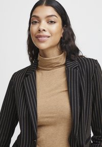 Black pinstriped blazer with notched lapels over a fitted tan turtleneck. Smooth fabric texture, tailored design, and prominent vertical stripes.
