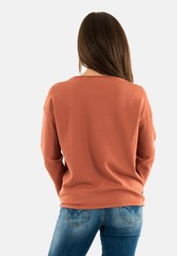 Donna con lunghi capelli castani, che indossa un maglione color ruggine e jeans blu, di spalle contro uno sfondo chiaro e semplice.