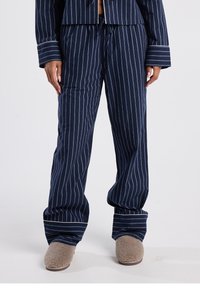 Navy blue pajama pants with white vertical stripes, relaxed fit, elastic waistband, and wide cuffs; paired with beige fuzzy slippers.