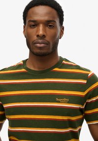 Grøn t-shirt med vandrette striber i gul, orange og rød. Har en rund halsudskæring og et lille "SuperDry Athletics" logo i orange.