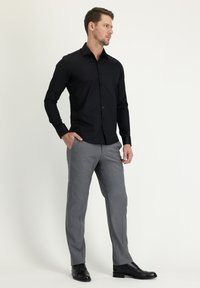 Camisa negra de manga larga con botones y cuello, combinada con pantalones grises claros y zapatos de vestir negros, de pie contra un fondo claro.