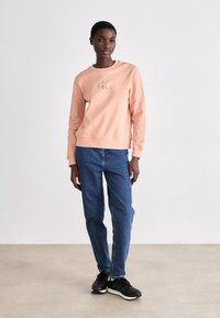 Sudadera rosa clara con escote redondo y logo bordado, combinada con jeans azul y zapatillas negras, sobre un fondo neutro.
