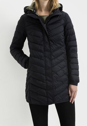 Femme portant une doudoune matelassée noire avec capuche, debout avec une main dans la poche et l'autre détendue le long du corps.