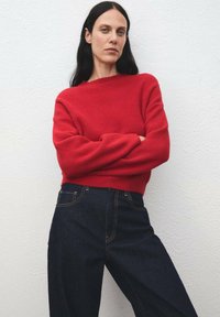 Donna con lunghi capelli scuri che indossa un maglione rosso e jeans blu scuri a vita alta, si trova con le braccia incrociate contro un muro bianco semplice.