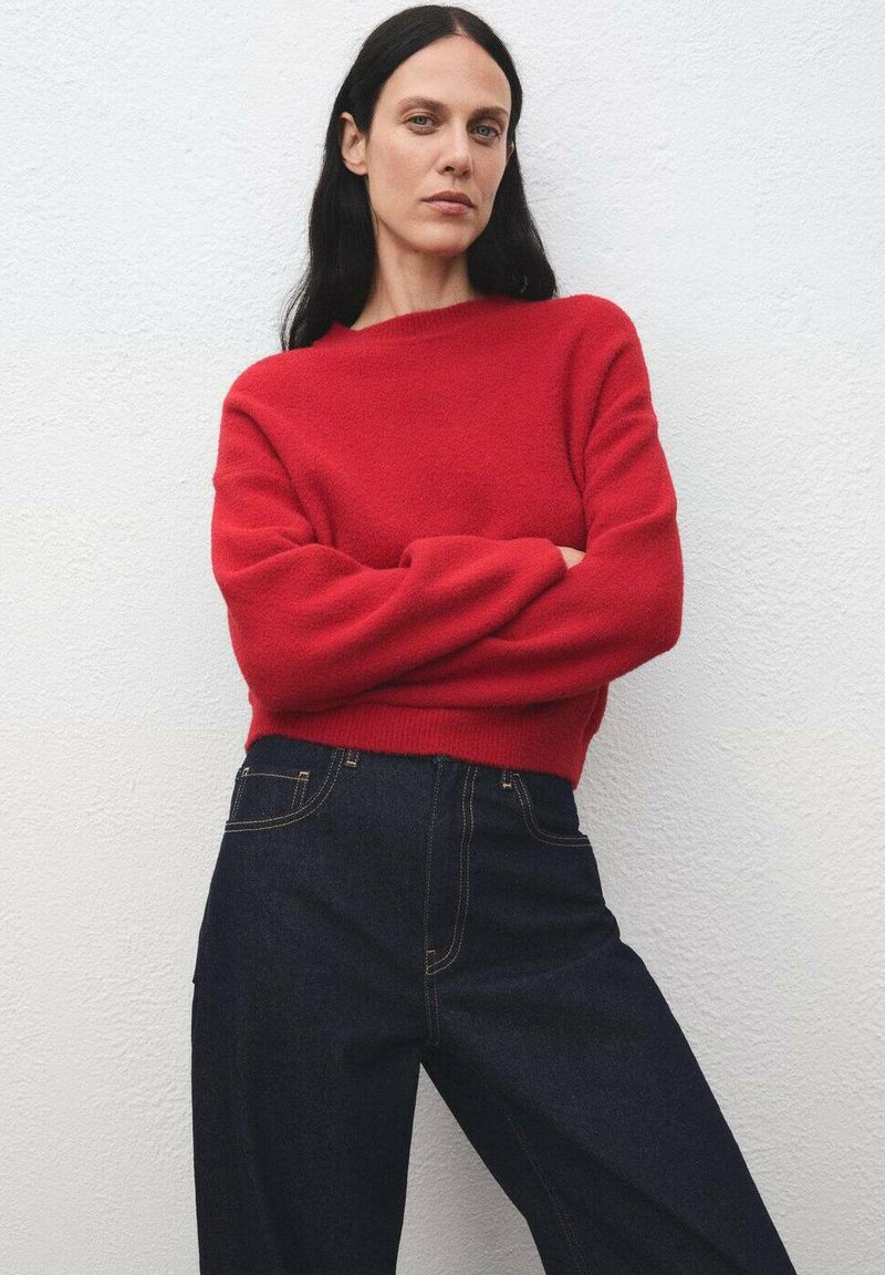 Donna con lunghi capelli scuri che indossa un maglione rosso e jeans blu scuri a vita alta, si trova con le braccia incrociate contro un muro bianco semplice.