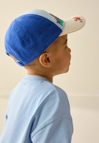 Baseballcap med blåt stof bagpå, cremefarvet frontpanel med grafik. Lys blå t-shirtkrave synlig, blød tekstur. Børneprofil.