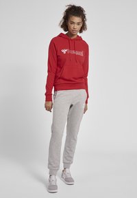 Czerwona bluza z kapturem z kieszenią kangurką z przodu oraz białym logo, w zestawieniu z jasno szarymi spodniami dresowymi z elastycznymi mankietami i białymi detalami, noszona z szarymi trampkami.