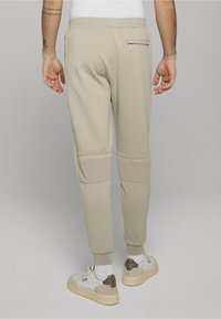 Béžové teplákové kalhoty s elastickým pasem, zúženými nohavicemi, bočními kapsami a texturovanými akcenty na kolenou. Nošeny s bílými teniskami.
