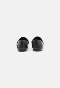 Zwarte leren loafers met een gladde afwerking, ronde neus en een lage hak. De achterkant is licht gebogen, wat het minimalistische ontwerp benadrukt.