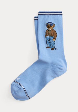 Lyseblå sok med en teddybjørn iført en blå kasket, brun jakke, stribet skjorte og jeans strikket på siden.