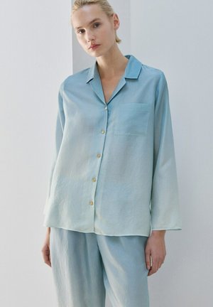 Femeie cu păr blond purtând un top cu nasturi din mătase bleu deschis și pantaloni largi asortați, stând în picioare pe un fundal simplu deschis.