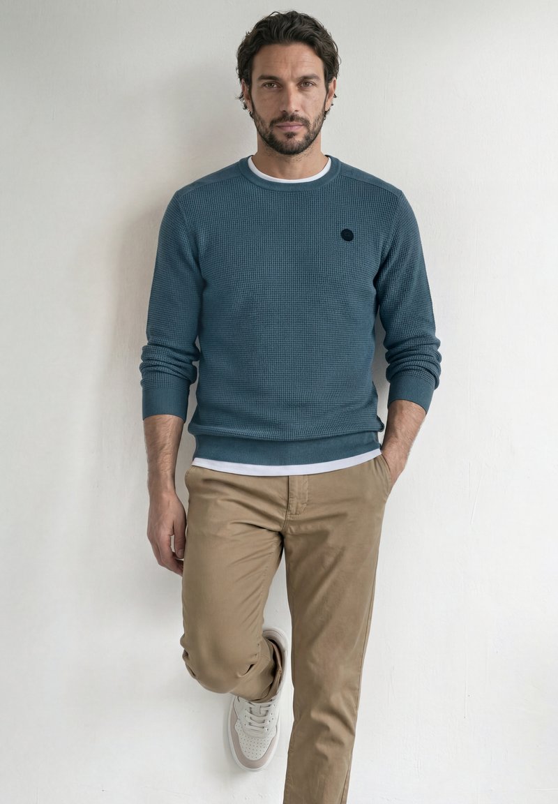 Homme aux cheveux foncés et à la barbe, portant un pull bleu, un pantalon beige et des baskets blanches, appuyé contre un mur blanc, une main dans la poche.