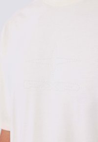 Camiseta de color crema claro con una textura suave, que presenta un sutil diseño de estrella en relieve y un patrón de puntos en el pecho.