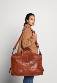 Sac à bandoulière en cuir marron avec deux poignées robustes, une sangle réglable et une poche zippée à l'avant. Texture lisse et forme rectangulaire.