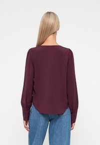 Pika lenge, tume lilla blouse ümmarguse kaelusega, kogutud õmblustega õlgadel ja nupudega accents varrukatel. Kombineeritud helesiniste teksadega.