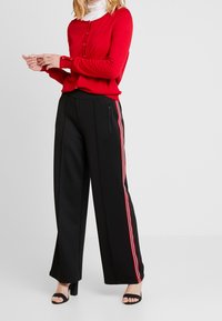 Röd kofta med knäppning, matchad med svarta vida byxor med röda sidostråk. Svart öppetåad klackskor fulländar outfiten.