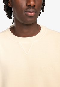 Beige sweatshirt met een geribbelde ronde hals en een subtiele V-vormige stikdetail. Geborduurd logo op de borst. Zachte, gestructureerde stof.