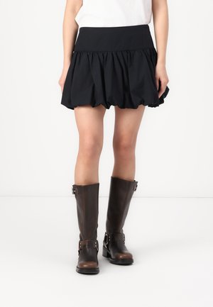 Woman wearing a black gathered mini skirt paired with knee-high dark brown leather boots against a plain background.