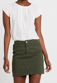 Vit kortärmad blus med veck, tillsammans med en olivgrön jeanskjol som har en diagonalsöm och framknäppning.