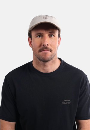 Casquette beige avec un design brodé, couronne ronde et visière courbée. Portée avec un T-shirt noir à manches courtes arborant un petit logo.