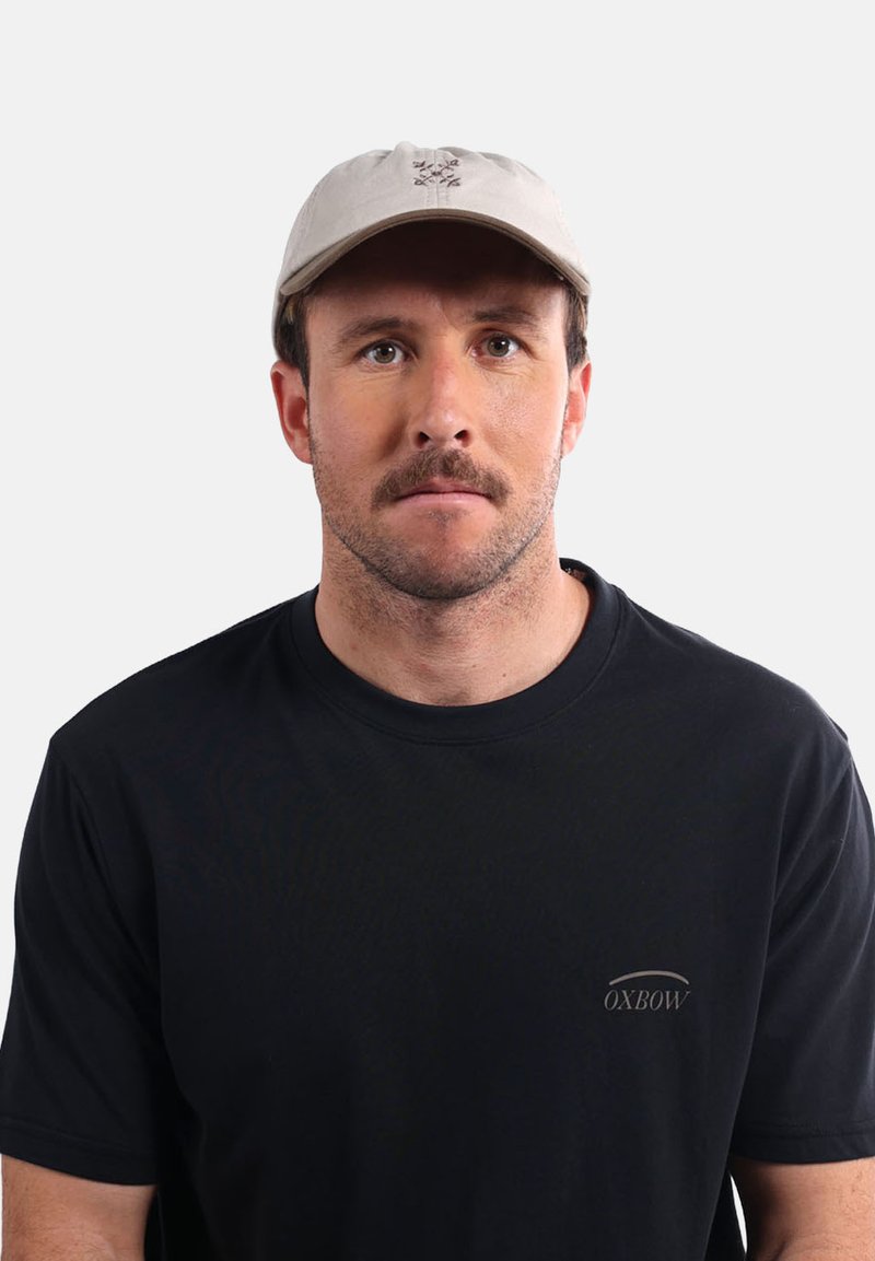 Casquette beige avec un design brodé, couronne ronde et visière courbée. Portée avec un T-shirt noir à manches courtes arborant un petit logo.